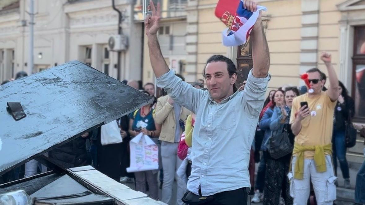David Martelo u Nišu ispred klavira sa zastavom Srbije u jednoj ruci, pokazujući tri prsta sa drugom
