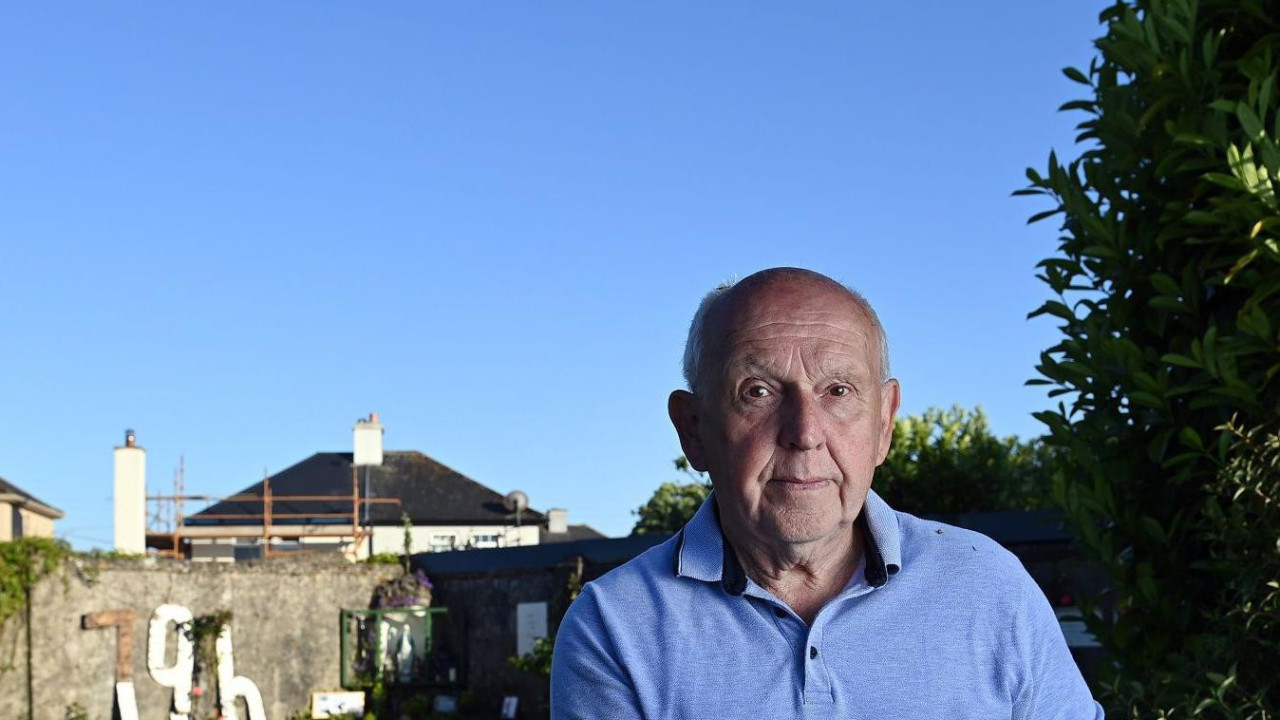 Čovek u plavoj polo majici stoji pored kamenog zida visokog do struka. U pozadini je parče travnjaka koje se proteže sve do visokog graničnog zida. Deo zida je prekriven bršljanom - na drugom delu se može videti broj 796 ispisan velikim, belim drvenim slovima. Čovek u prvom planu je stariji, ćelav i drži ruke na niskom zidu. Dan je i nebo je vedro plavo.