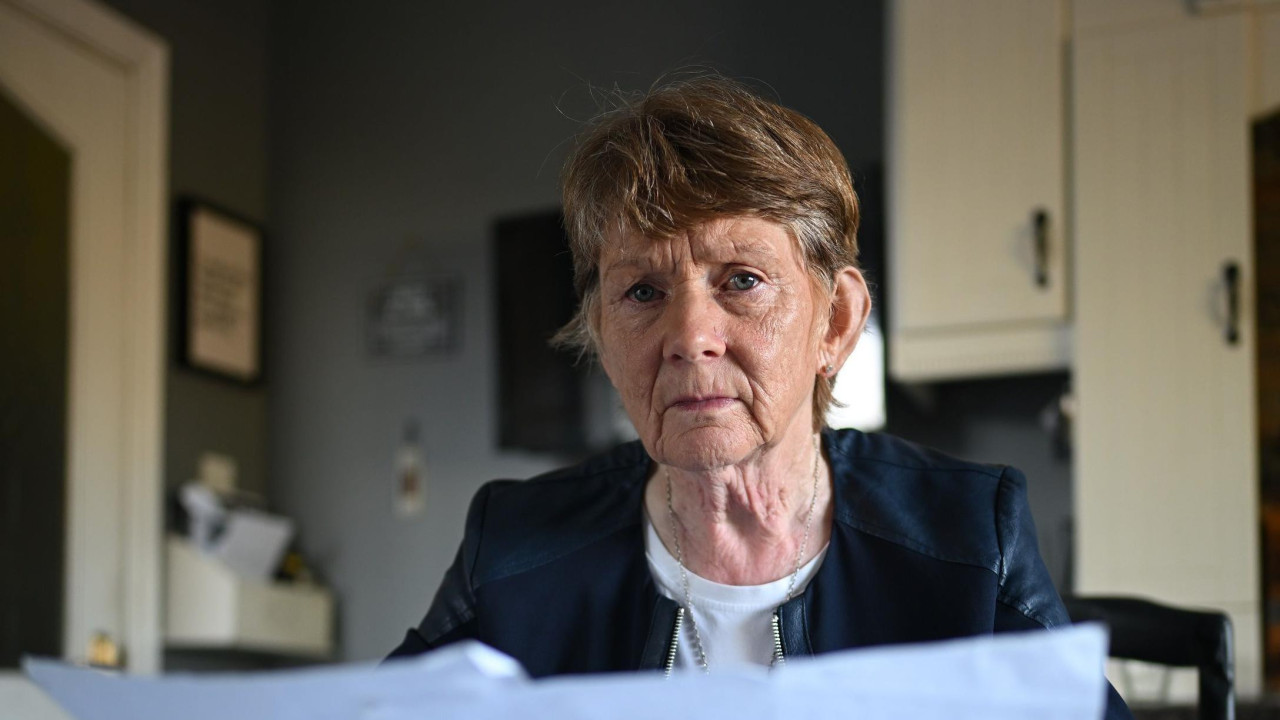 Starija žena sedi za kuhinjskim stolom. U svakoj ruci drži list papira. Na stolu ispred nje je još dokumenata i listova papira. Ima kratku smeđu, sedu kosu i nosi belu majicu i plavu jaknu. U pozadini se vide kuhinjski ormarići i kuhinjska vrata sa dva zastakljena prozora.