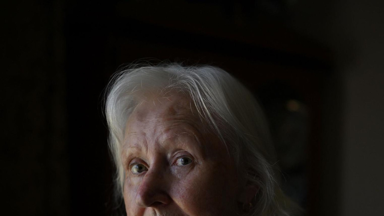 Starija žena sa srednje dugačkom sedom kosom stoji u dnevnoj sobi. Vidimo joj glavu i ramena. Nosi tamnu jaknu i šareni svileni šal. U pozadini, koja je van fokusa, vidimo sliku koja visi na zidu dnevne sobe i ormar.