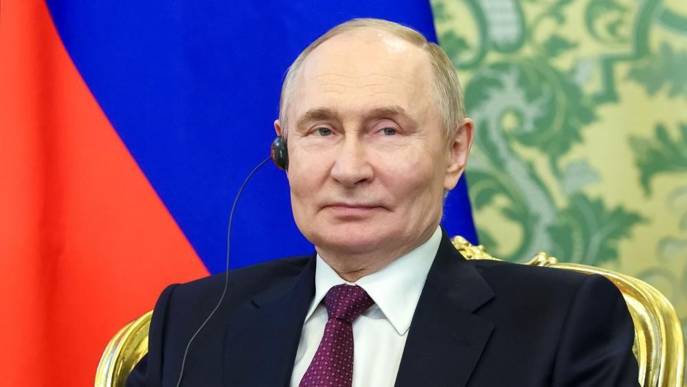 A headshot portrait of Vladimir Putin smiling slightly against the red and blue backdrop of a flag